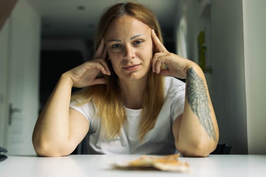 a girl of European appearance in a white T-shirt sits at a white table and looks straight into the camera, hands holding her head on her left arm tattoo .blurred background. High quality photo