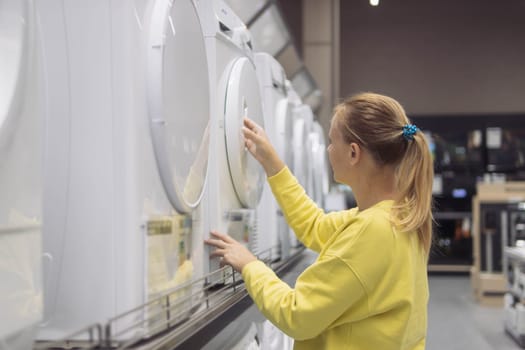 a girl with blond hair tied in a ponytail in a yellow sweater, looking at a washing machine in a store. Close-up of her face and hands, the girl is not looking at the camera. High quality photo