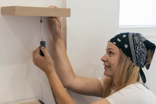 a girl of European appearance in a white T-shirt on her head with a black bandana, twists screws into a shelf on the wall with a screwdriver. there is a place for an inscription. High quality photo