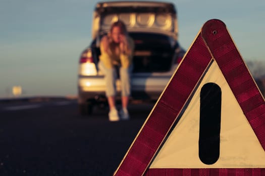 a girl in casual clothes sits in the trunk, in the foreground is an emergency stop sign close-up and in focus, the girl is blurred in the background. Car breakdown on the highway. High quality photo