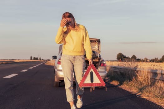 a girl of European appearance with blond hair, goes to put an emergency stop sign on the highway near a broken car. A car breakdown on the road in weight,a place for an inscription. High quality photo