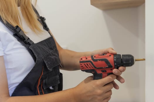 a beautiful girl of European appearance in a black bandana, white t-shirt and in a special suit drills a hole in a white wall with a drill. The concept of equality and work. High quality photo