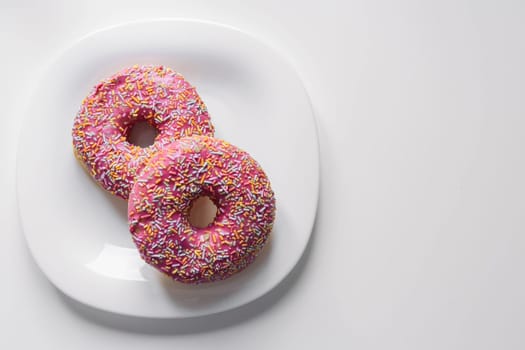 two donuts with a pink icing and colored sprinkles on a white plate lie on a white table, on the right there is a place for an inscription. High quality photo