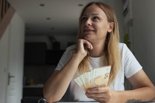 a girl with blond hair sits indoors at a white table with her hands folded and the table is thoughtful looks to the side, in her hands the girl holds euros of 50 salaries. High quality photo