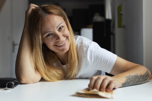 a girl with blond hair sits indoors at a white table cheerful holds her hand on the salt on the money with the other hand her head on the left hand of the girl tattoo. High quality photo