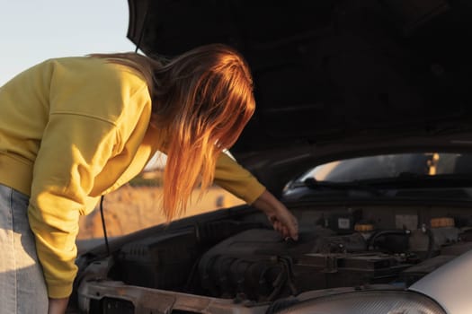 a girl with blond hair, stands with the hood of the car open and looks at what has broken. A car breakdown on the road. there is a place for an inscription on the right. High quality photo