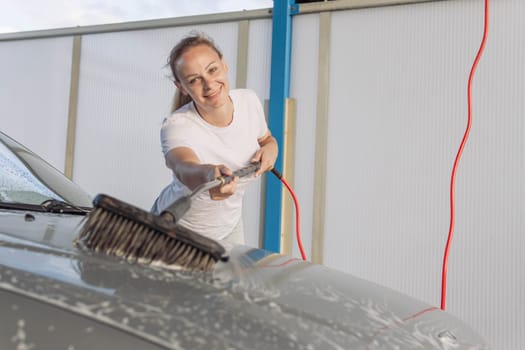 A young woman washing a car smiles. A girl in a white T-shirt with blond hair braided in a ponytail has a place for an inscription. High quality photo