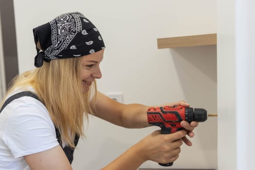 a beautiful girl of European appearance in a black bandana, white t-shirt and in a special suit drills a hole in a white wall with a drill. The concept of equality and work. High quality photo