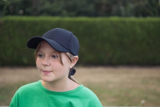 a teenage girl of European appearance was dreaming in a black cap, in a green T-shirt, standing in a park around green grass on the right there is a place for an inscription. High quality photo