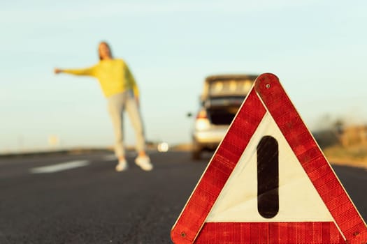 in the foreground, an emergency stop sign in focus, in the background, a blurred girl in a yellow jacket stands with her hand raised, stops the car for roadside assistance. High quality photo