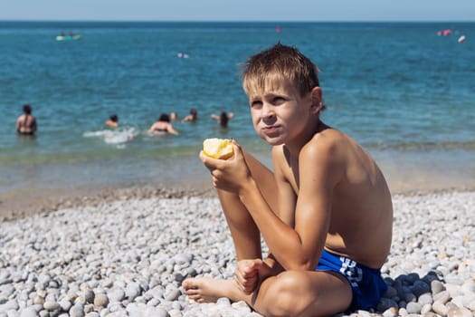 a tanned blond boy with blond hair is sitting on the beach eating an apple near the sea in blue shorts. High quality photo