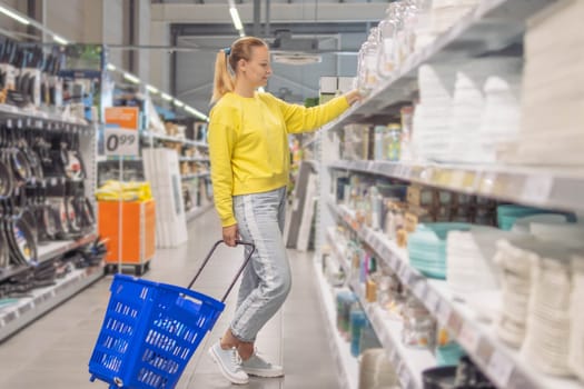 Cheerful girl makes purchases in a dishware store, chooses beautiful dishes for home. High quality photo a girl with blond hair of European appearance in jeans and a yellow sweater stands