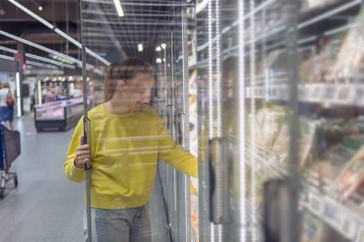 a woman with blond hair tied in a ponytail in a yellow sweater and jeans, in a store in a refrigerated display case chooses products for the home. The concept of home food. High quality photo