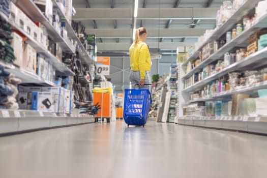 a girl with blond hair tied in a ponytail in jeans and a yellow sweater stands between the counters in a store with a blue plastic cart turned her back. High quality photo