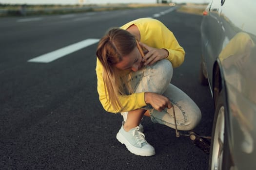 a girl in a yellow sweater and jeans of European appearance sits near the rear punctured wheel in her hands holds a key for the wheels and speaks on the phone how to make a wheel. High quality photo