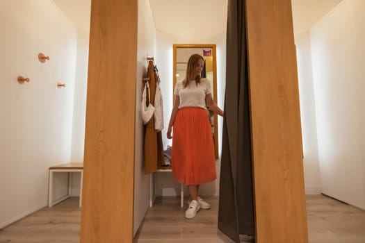 a girl trying on a white t-shirt and an orange skirt in a booth looks in the mirror. brown and white fitting booths in a things store. there is a place for an inscription. High quality photo