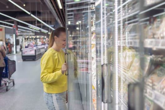 a woman with blond hair tied in a ponytail in a yellow sweater and jeans, in a store in a refrigerated display case chooses products for the home. The concept of home food. High quality photo