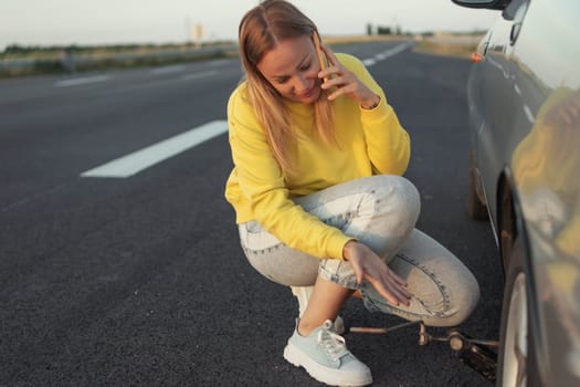 a girl in a yellow sweater and jeans of European appearance sits near the rear punctured wheel in her hands holds a key for the wheels and speaks on the phone how to make a wheel. High quality photo