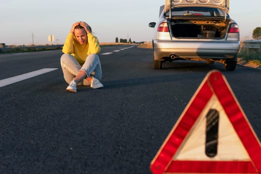 a woman in a yellow sweater and jeans of European appearance sits on the pavement near a broken car, upset, worries next to it is a white-red emergency stop sign. High quality photo