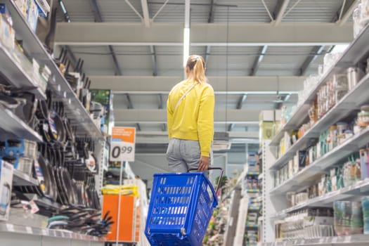 a girl with blond hair tied in a ponytail in jeans and a yellow jacket walks between the shelves in a store with a blue plastic cart turned close-up. High quality photo