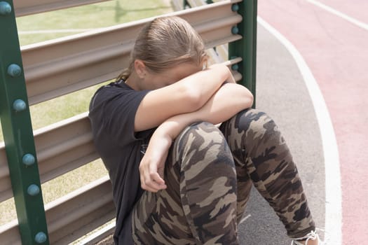 a teenage girl with blond hair sits by a fence near a football field, covering her face with her hands. in white sneakers, a dark T-shirt and green leggings. High quality photo