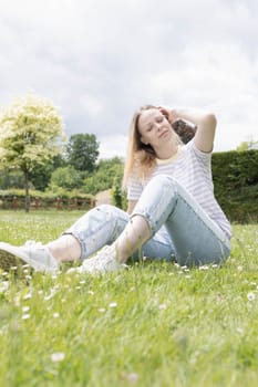 a girl with blond hair of European appearance sits in a park on green grass, in jeans and a white T-shirt on her left arm a tattoo in the shape of a wing. High quality photo