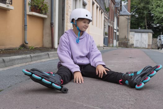 a teenage girl of European appearance in a purple sweater sits on the ground in roller skates, learns to roller skate. High quality photo child in a white safety helmet