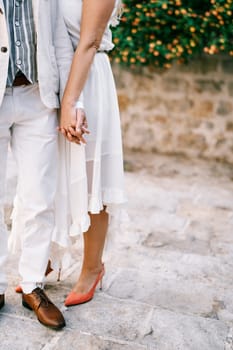 Bride and groom are standing holding hands on the paving stones in the garden. Cropped. Faceless. High quality photo