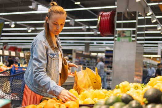 girl of fair appearance with tied hair, in a skirt and a denim jacket, stands near the counter with yellow pepper and chooses it,The concept of healthy eating and vegetarianism. High quality photo