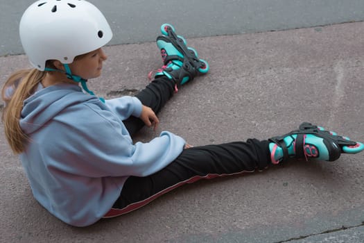 a girl with blond hair sits on the pavement in roller skates, takes a break from studying rollerblading. A teenager in black pants and a blue jacket. High quality photo child in a white safety helmet