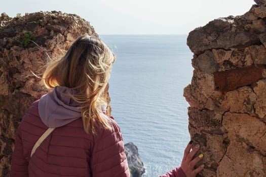 a girl in a pink jacket with blond hair stands backwards and looks at the sea from a height, a beautiful landscape. High quality photo