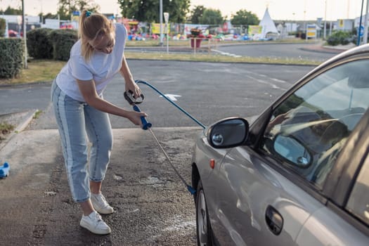 girl of European appearance in a white t-shirt and jeans with blond hair tied in a ponytail, washes a gray car with water outdoors at the sink. The concept of work and cleanliness. High quality photo