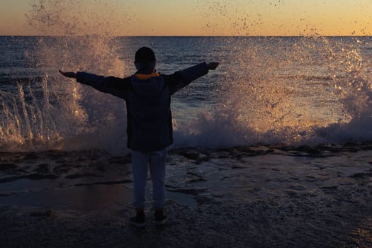 the child is standing on the seashore hands up, photo from the back, the waves are raging at sunset, the background. High quality photo