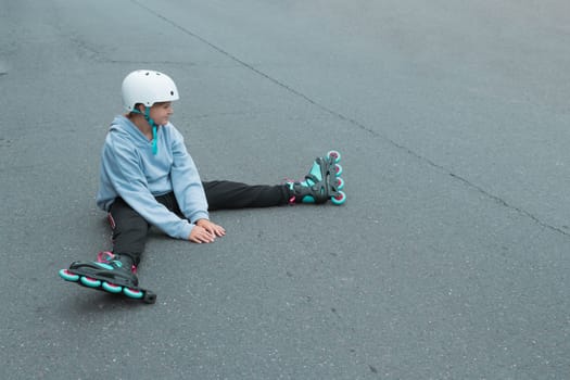 a teenage girl of European appearance in a sweater sits on the ground in roller skates, learns to roller skate. High quality photo child in a white safety helmet