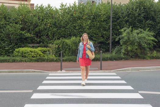 happy girl with blond hair in a red dress, denim jacket and with a bag on her shoulder crosses the road on a pedestrian crossing front view back beautiful green trees. High quality photo