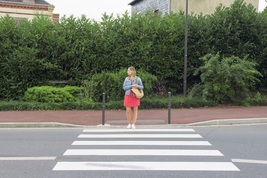 A girl in a red dress and a denim jacket stands at a pedestrian crossing. pedestrian crossing on the road for safety when people cross the street, Traffic rules concept. High quality photo