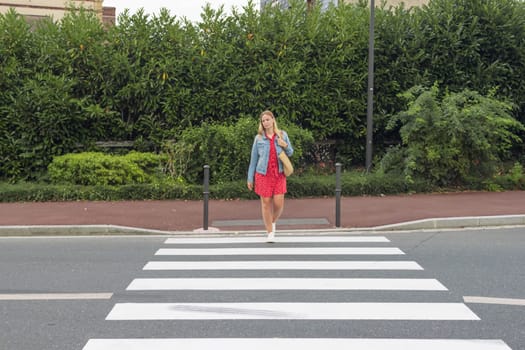 A girl with fair hair in a red dress and a denim jacket with a bag on her shoulder crosses the road at a pedestrian crossing.a pedestrian crossing on the road for safety. High quality photo