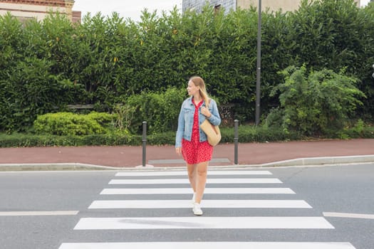 A girl in a red dress and a denim jacket stands at a pedestrian crossing. pedestrian crossing on the road for safety when people cross the street, Traffic rules concept. High quality photo