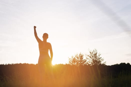 girl in a fitness suit stands with her back to the camera at sunset with her hand raised up after a workout in the fresh air, motivational girl silhouette place for an inscription. High quality photo