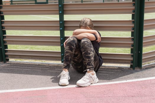 a teenage girl with blond hair sits by a fence near a football field, covering her face with her hands. in white sneakers, a dark T-shirt and green leggings. High quality photo