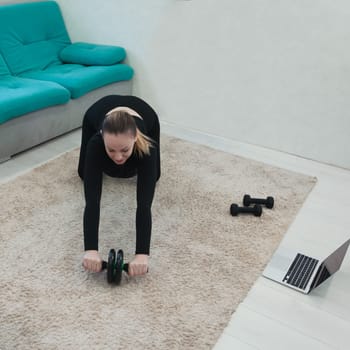 A young girl of European appearance goes in for sports online in a laptop in a room during quarantine in sportswear. Get on your knees and use the ab roller to stretch your body. Home training.