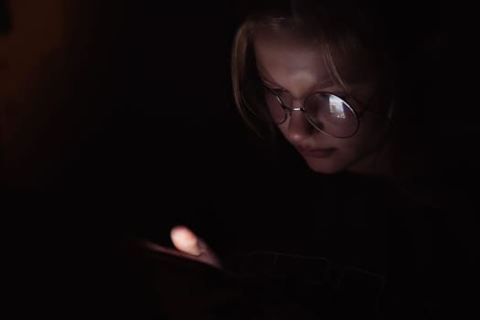 a teenage girl of European appearance in glasses for vision, sits in a dark room looking at the phone, the light falls on the girl's face, there is a place for an inscription. High quality photo