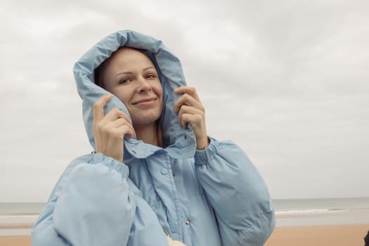 a girl of European appearance stands on the seashore in a blue jacket in a hood and smiles, looks into the camera. High quality photo