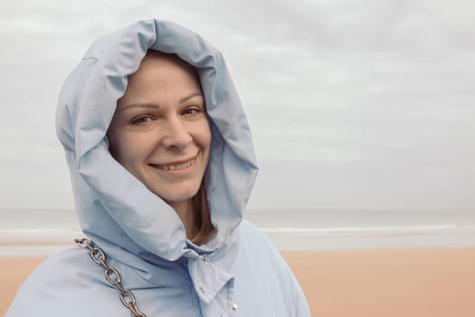 a girl of European appearance stands on the seashore in a blue jacket in a hood and smiles, looks into the camera. High quality photo