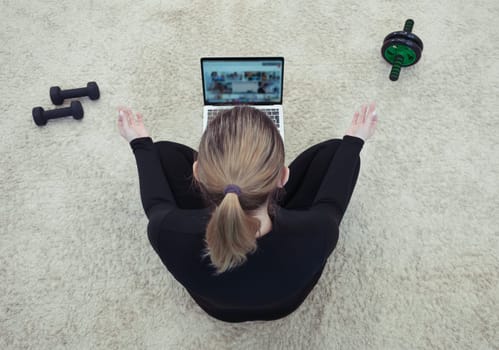 Rear view, Athletic healthy woman sitting on mat in lotus position, wearing sports black uniform, watching online yoga classes, meditating, breathing exercises on laptop. sports equipment is nearby.