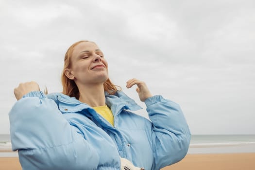 a girl of European appearance with blond hair stands on the seashore in a blue jacket and smiles with her eyes closed. High quality photo
