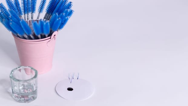 On a white background, a layout with materials for eyelash and eyebrow extensions. On the left, on a white background, there is a pink bucket with blue brushes for eyelashes