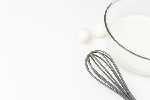 a glass transparent bowl with milk on a white table, next to a whisk for beating and eggs. close-up on a white table Culinary hobby at home - baking: cakes, muffins. High quality photo