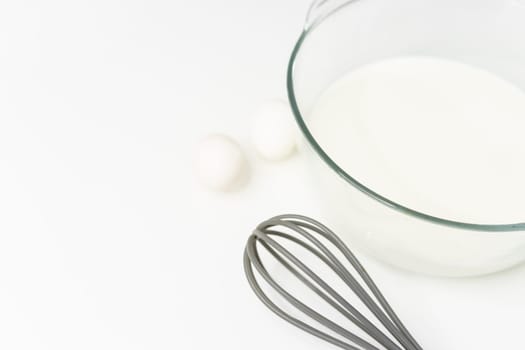 a glass transparent bowl with milk on a white table, next to a whisk for beating and eggs. close-up on a white table Culinary hobby at home - baking: cakes, muffins. High quality photo