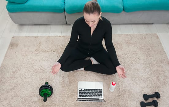 Sporty healthy calm woman sitting on mat in lotus position,wearing sporty black uniform,watching online yoga class,meditate,do breathing exercises on laptop.White bottle with water is standing nearby.